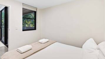 A minimalist bedroom with a single bed, a window with a view of greenery, and a door leading to an outdoor area.