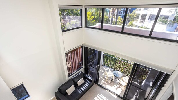 A modern living room with a black leather sofa, a round wooden coffee table, and a large sliding glass door leading to an outdoor patio area. - The Aubrey Boutique Apartments (Surfers Paradise)