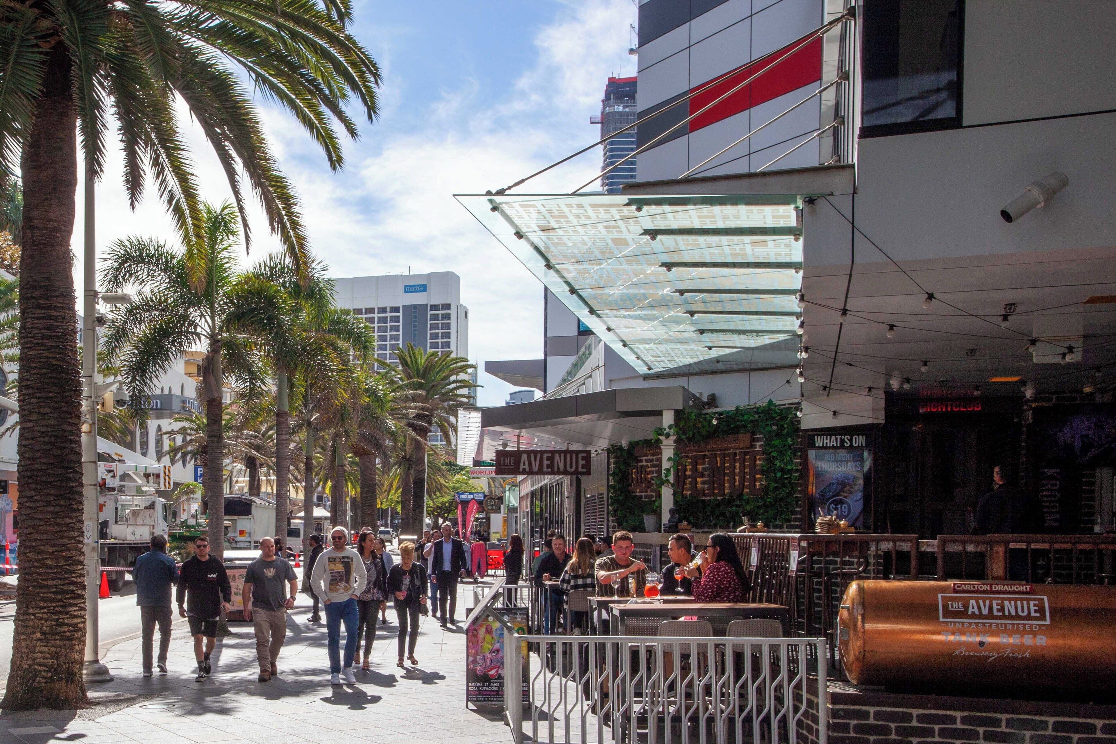 Une rue animée en ville, bordée de palmiers, avec un bar appelé «The Avenue» et des gens qui se promènent.