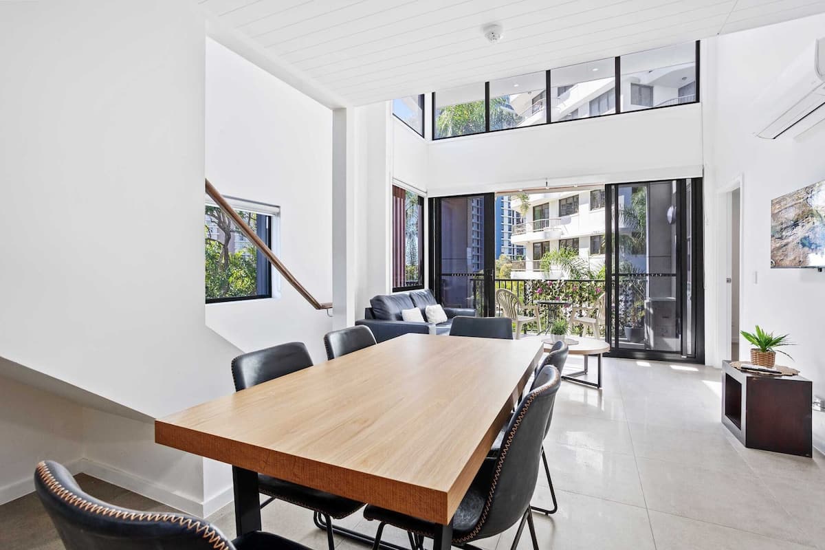 A modern dining area with a wooden table and black chairs, large windows, and a view of the outside balcony.
