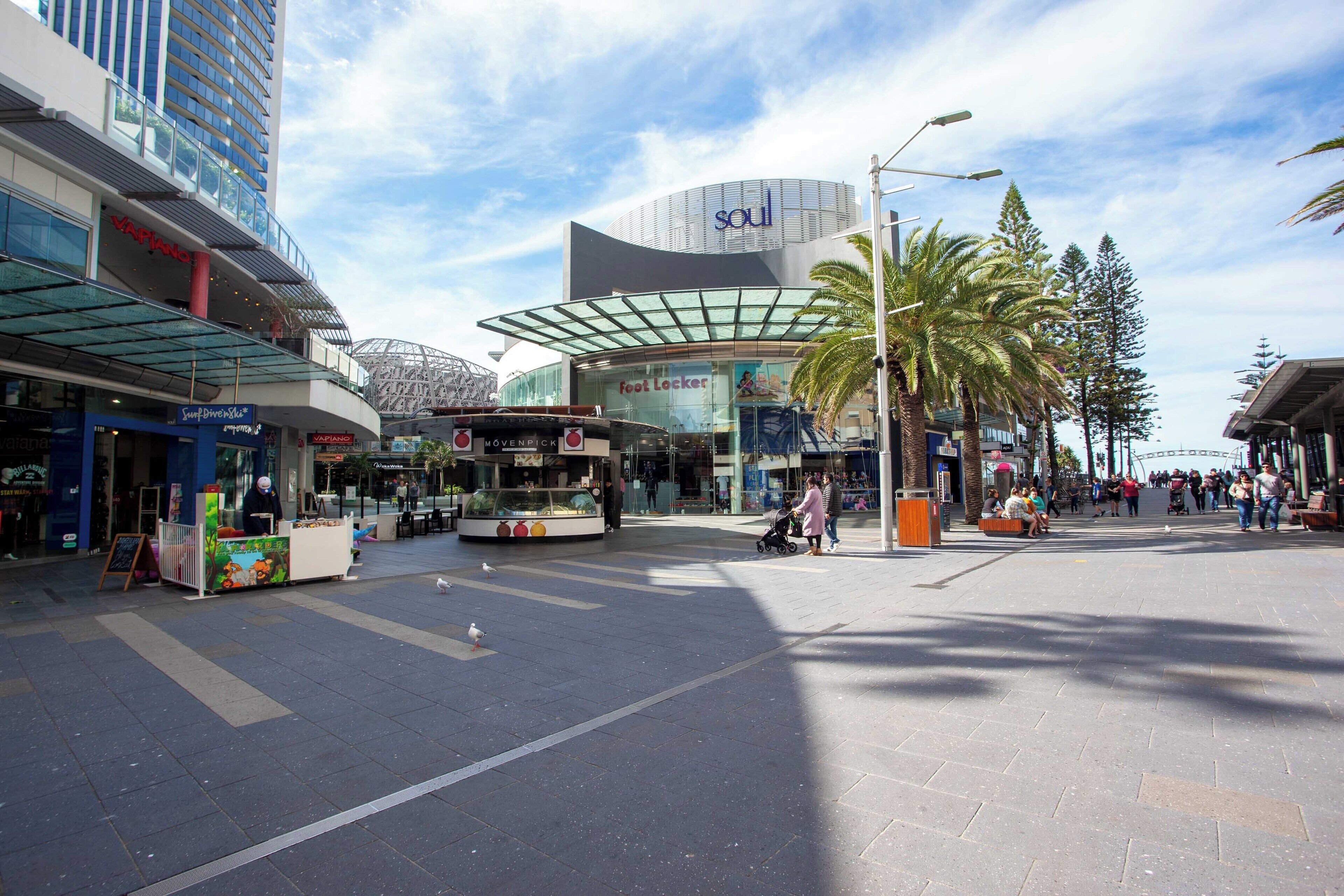 Une zone piétonne avec des boutiques, un kiosque à nourriture et un grand bâtiment aux lignes courbes, doté d’une façade en verre.