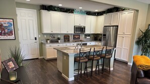 Una cocina con armarios blancos, una isla central con taburetes y electrodomésticos como refrigerador y microondas.