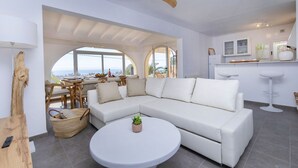 Een moderne woonkamer met een witte modulaire bank, een ronde salontafel met een plant, een eethoek met uitzicht, en een keuken met witte kasten.