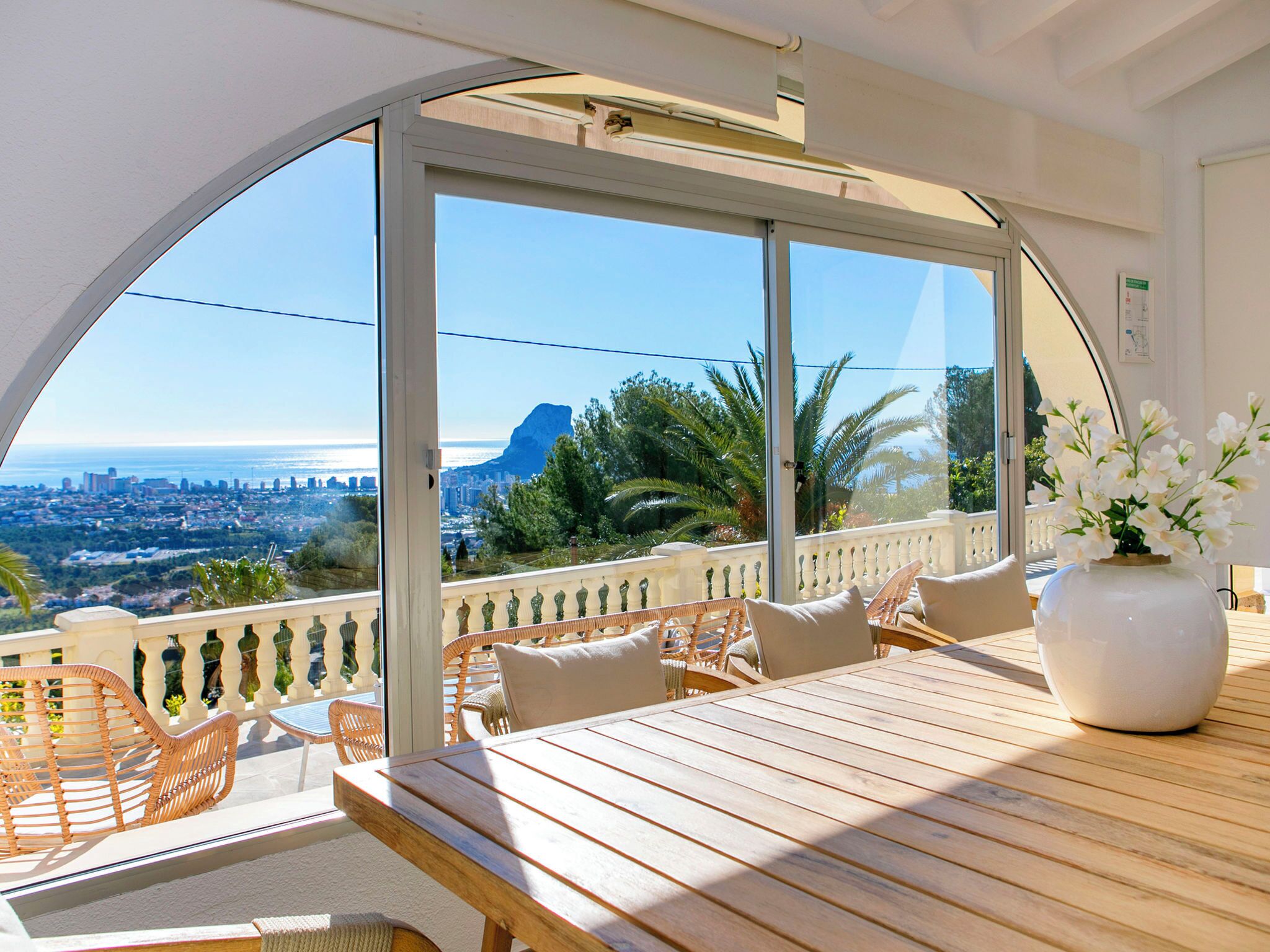 A balcony with a view of the city and the sea, furnished with wicker chairs and a wooden table.