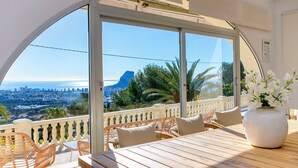Een balkon met uitzicht op de stad en de zee, voorzien van rieten stoelen en een houten tafel.