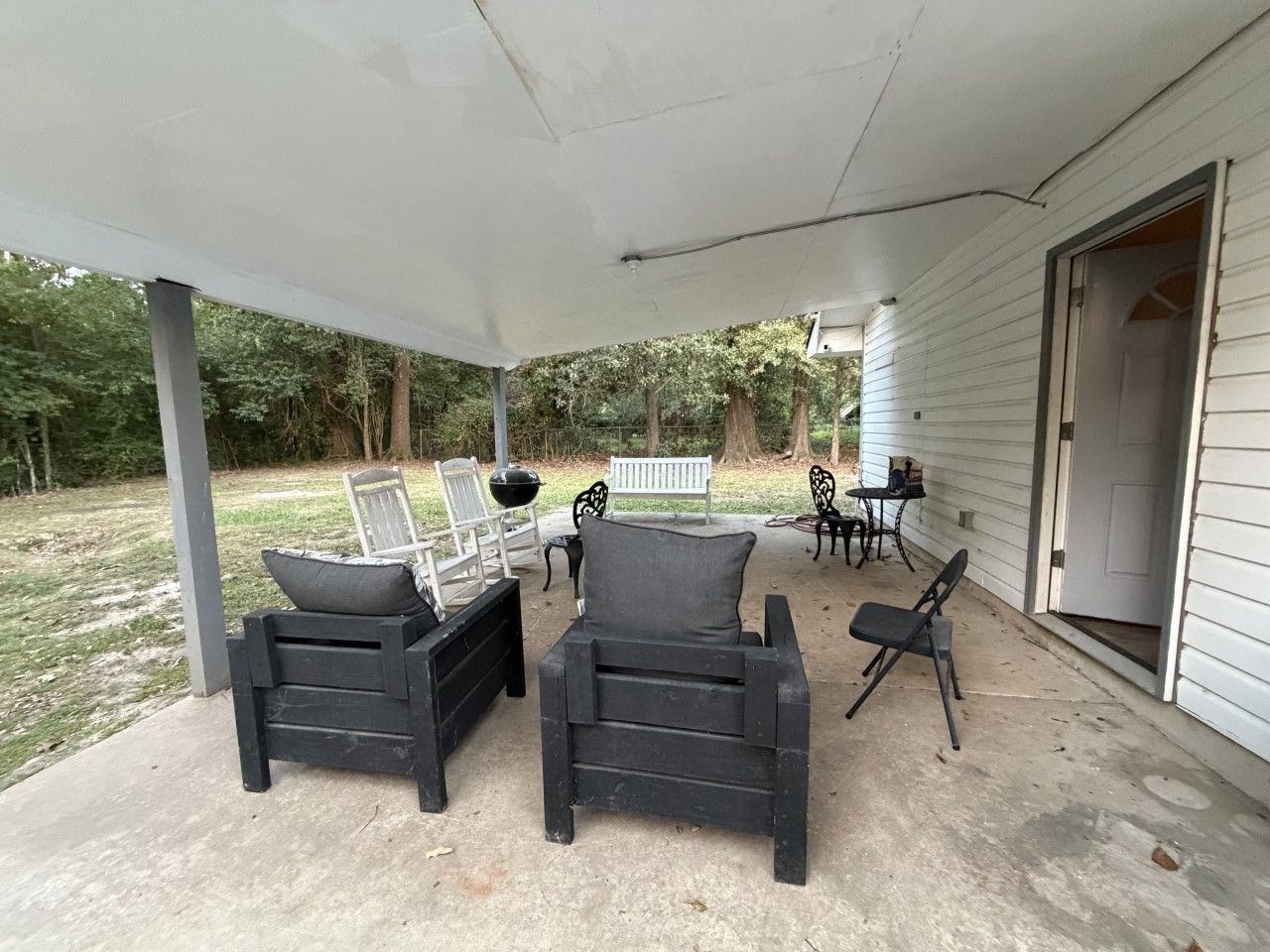 An outdoor covered patio with two black chairs and a bench, a white door, and a small table with a chair.