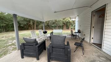 An outdoor covered patio with two black chairs and a bench, a white door, and a small table with a chair.