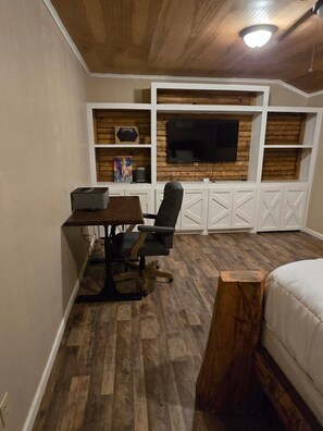 A room with a wooden ceiling, a desk with a chair, a bed, and built-in shelving with a television.
