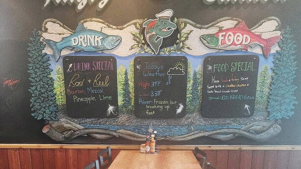 A restaurant interior with a long wooden table, chairs, and a chalkboard menu displaying drink and food specials.