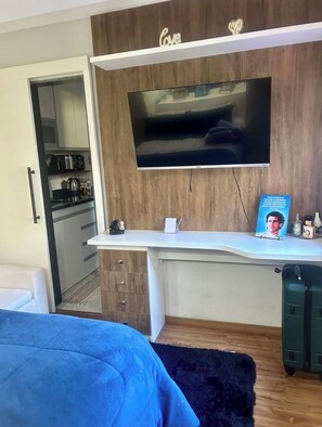 A modern bedroom with a TV mounted on the wall, a desk with a chair, and a small kitchenette in the background.