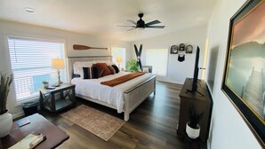A bedroom with a bed, bedside tables, a desk, a chair, a ceiling fan, and a large window with blinds.