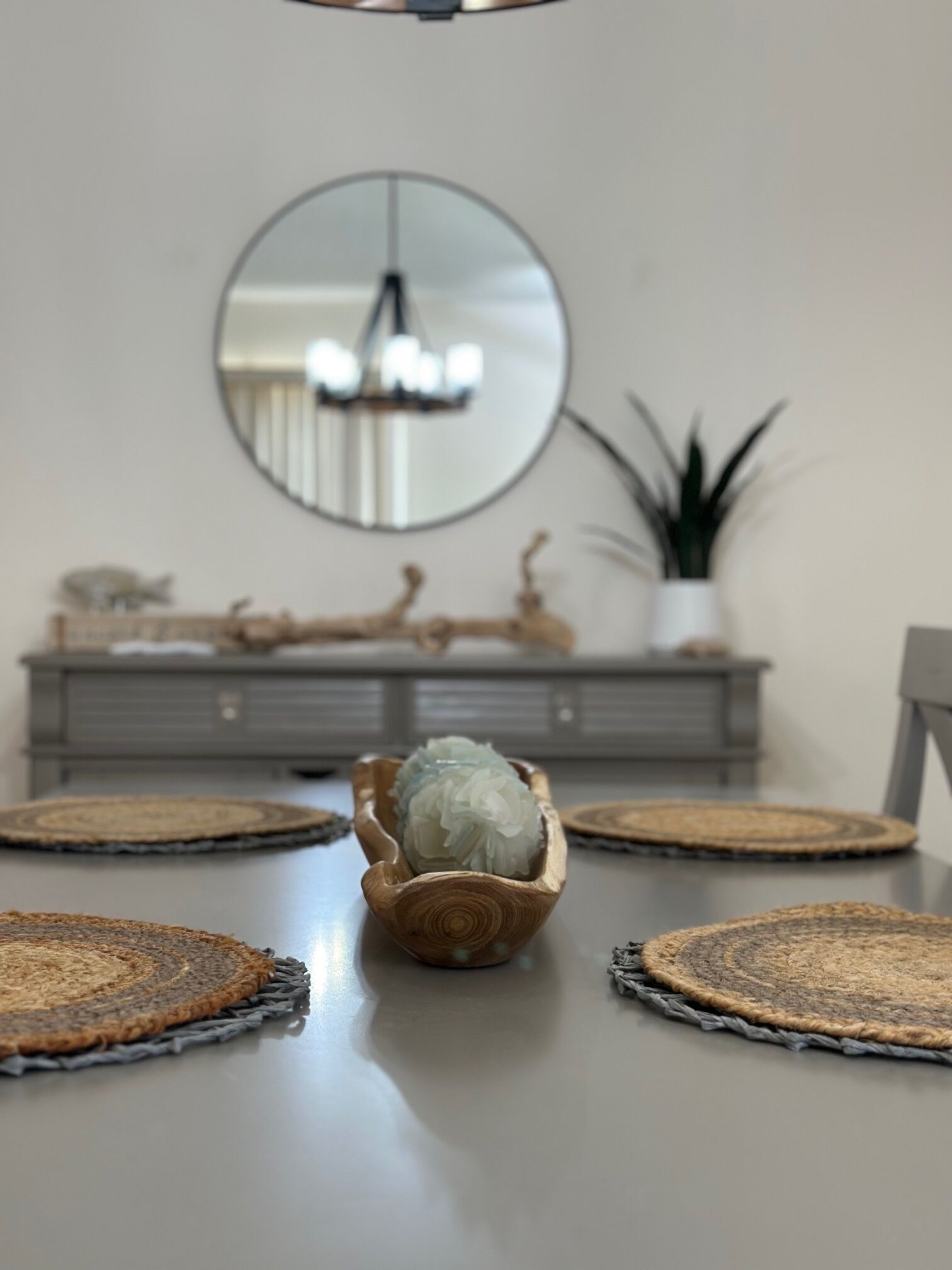 A dining table with a central decorative bowl, surrounded by woven placemats, a grey console table with a mirror and decorative items, and a potted plant.