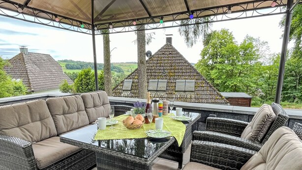 Une terrasse extérieure avec des meubles en osier, une table recouverte d’une nappe verte, et une vue sur des maisons en arrière-plan.