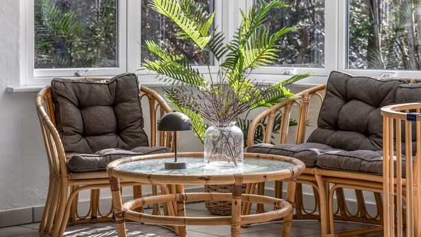 Une pièce avec une table ronde en verre, entourée de chaises en osier dotées de coussins gris. Un vase transparent contenant des feuilles vertes est posé sur la table.