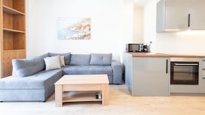 Een moderne woonkamer met een grijze modulaire bank, een houten salontafel, en een keukenhoek met grijze kastjes en een inbouwoven.