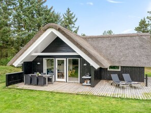Ein modernes Haus mit einem Strohdach, einer Terrasse mit Gartenmöbeln und einer Schiebetür zu einem Innenraum.