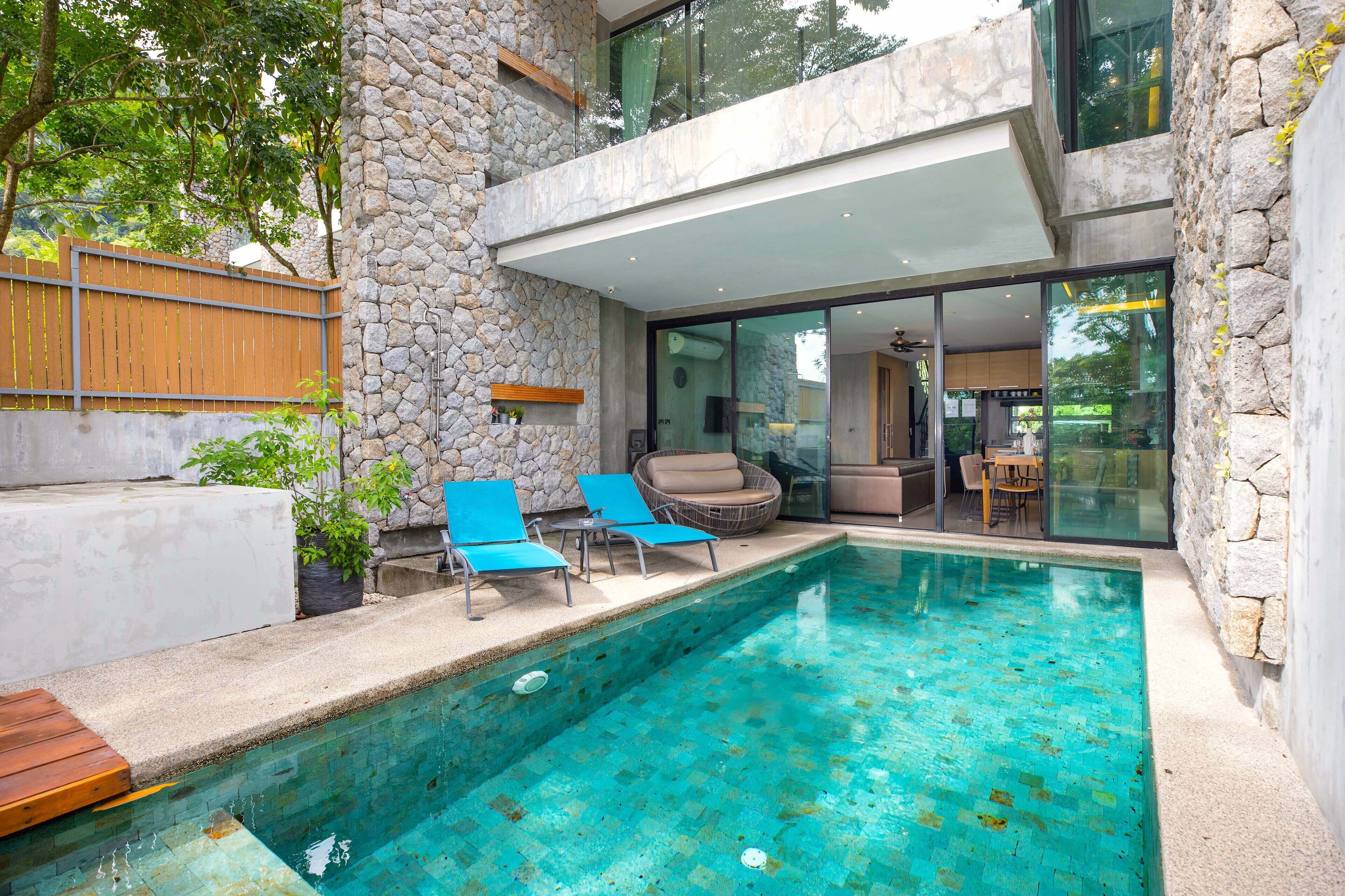 A modern outdoor pool area with stone walls, two lounge chairs, and a glass-enclosed patio.