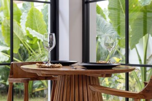 A wooden dining table with two wine glasses and plates, set against a backdrop of large green leaves through a window.