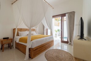 A bedroom with a wooden bed, white bedding, a mosquito net, a bedside table, a TV, and a view of the outside.