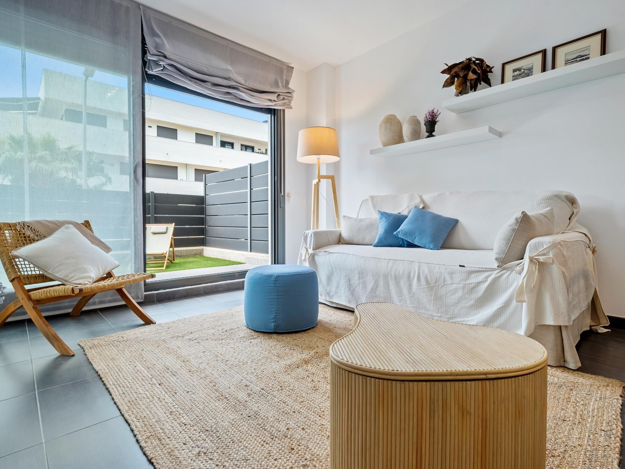 Een moderne woonkamer met een bank, een blauwe poef, een rieten fauteuil, een lamp en uitzicht op een gebouw door een groot raam.