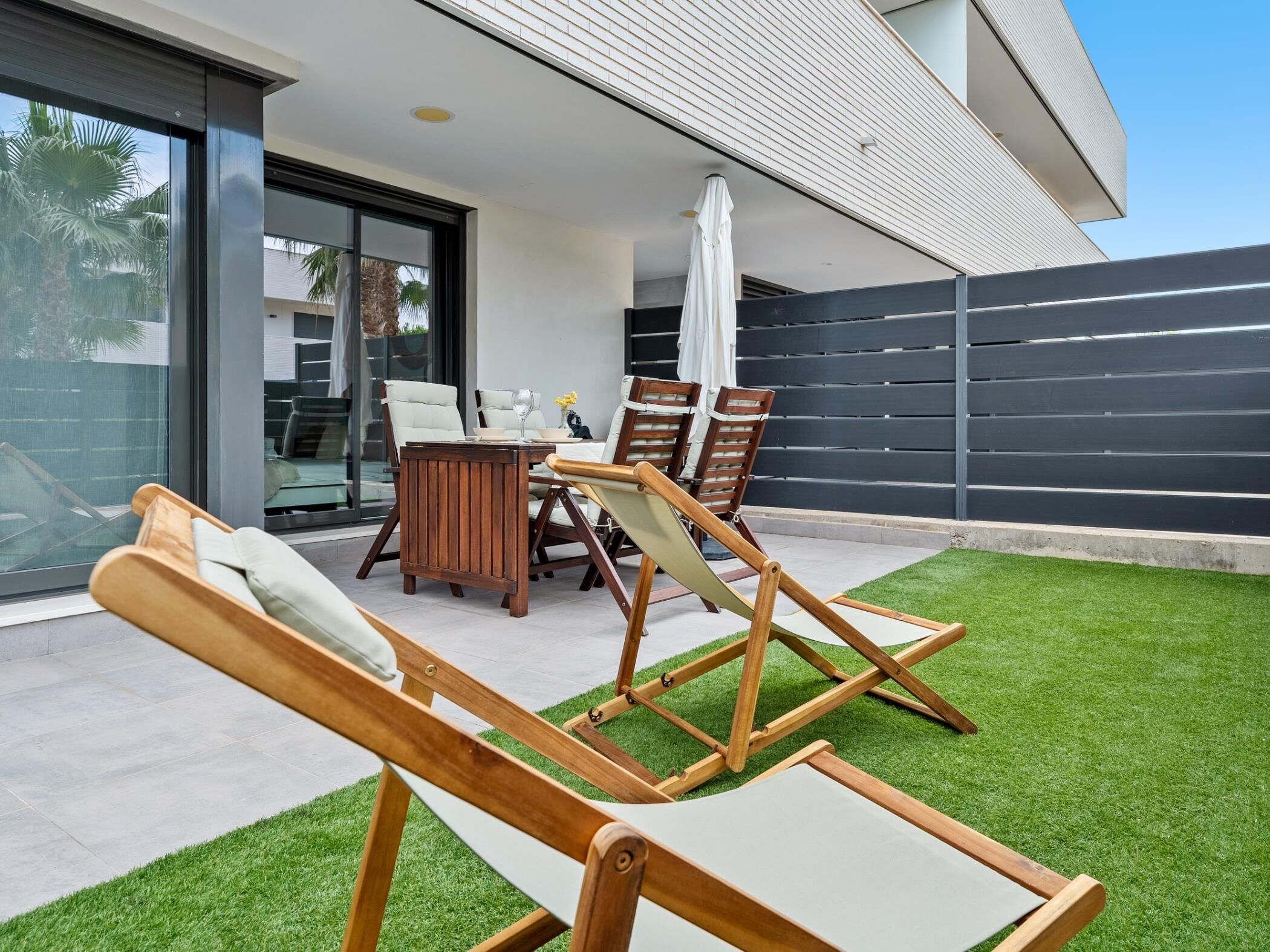 Una moderna veranda esterna con tavolo da pranzo, sedie e una zona relax con poltrone prendisole in legno.