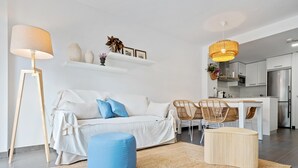 Een moderne woonkamer met een bank, voetenbankje en stoelen, een keuken op de achtergrond en een grote vloerlamp.