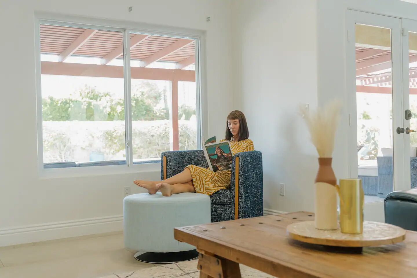 A person sitting on a blue chair in a room with a large window, a wooden table, and a vase with yellow flowers.