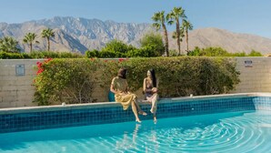 Una piscina amb dues persones assegudes al costat, envoltada de vegetació i muntanyes al fons.