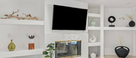 A modern living room with a flat-screen TV mounted above a fireplace, white shelves with decorative items, and a potted plant.