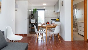 A modern kitchen with white cabinets, a dining area with a wooden table and white chairs, and a grey sofa in a living room.