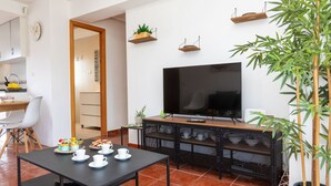 A modern living room with a TV, a dining table with cups and a plant, and a kitchen in the background.