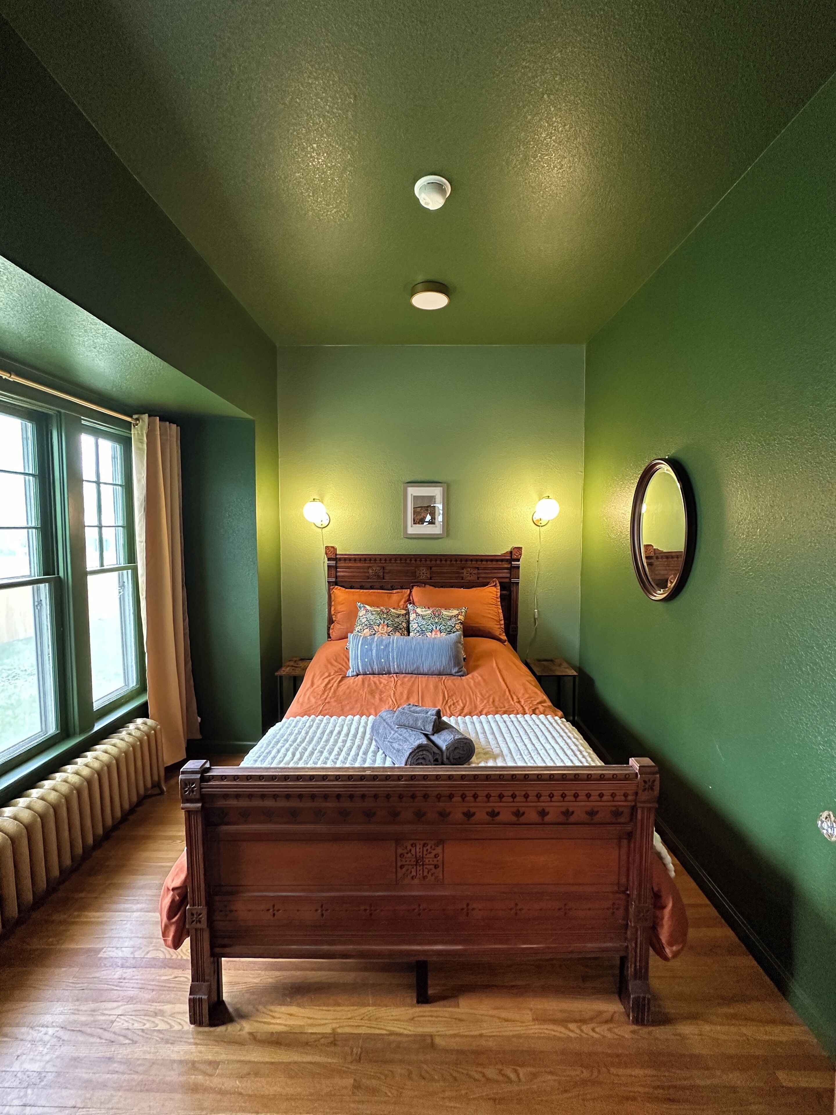 A bedroom with a wooden bed, green walls, a window with curtains, a radiator, and a ceiling light.