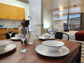 A dining area with a wooden table set for two, featuring white plates, clear wine glasses, and a vase with white flowers. - U Suites on Egmont (Wellington)