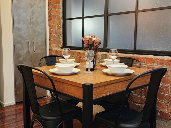 A dining table set with white bowls and wine glasses, surrounded by black chairs, in a room with brick walls and a large window. - U Suites on Egmont (Wellington)
