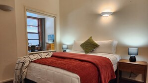 Ein ordentlich bezogenes Bett mit roter Decke, zwei Nachttischlampen und einem Fenster mit Jalousien.