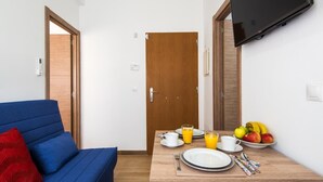 A compact living space with a blue sofa, a wooden dining table set with plates, cups, and a bowl of fruit, and a TV mounted on the wall. - Cozy Apartment on the Beach, ideal for couples (FUENGIROLA)