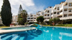 A swimming pool with a clear blue water surface, surrounded by white buildings with balconies and a palm tree. - Coral Island, Mediterranean Duplex Penthouse (BENALMADENA COSTA)