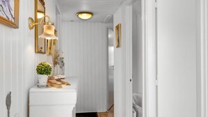 Un couloir aux murs blancs, avec un radiateur blanc, un miroir encadré d’or et un luminaire fixé au mur.