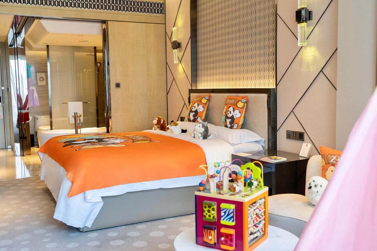 Habitación de hotel moderna con una cama grande, una mesita de noche con lámpara, un escritorio pequeño y un caja de almacenamiento de juguetes de colores.