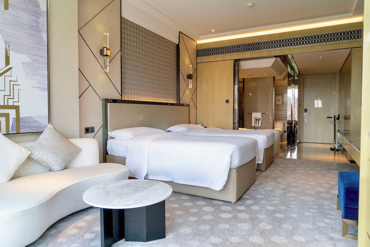 A modern hotel room with a large bed, a sofa, and a small table. There is a mirror and a door leading to another room.