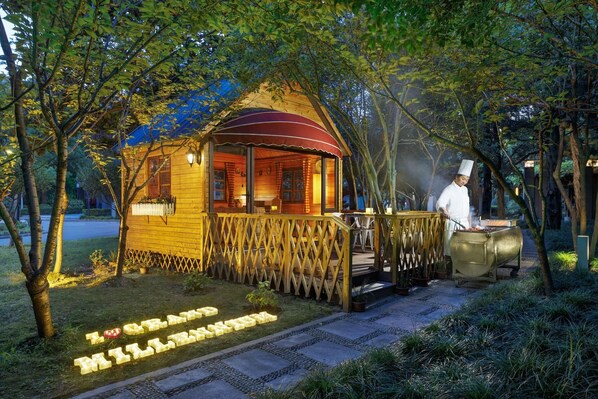 A wooden cabin with a covered porch, a barbecue grill, and a pathway leading to the cabin. - Grand Millennium Shanghai Hongqiao (Shanghai)