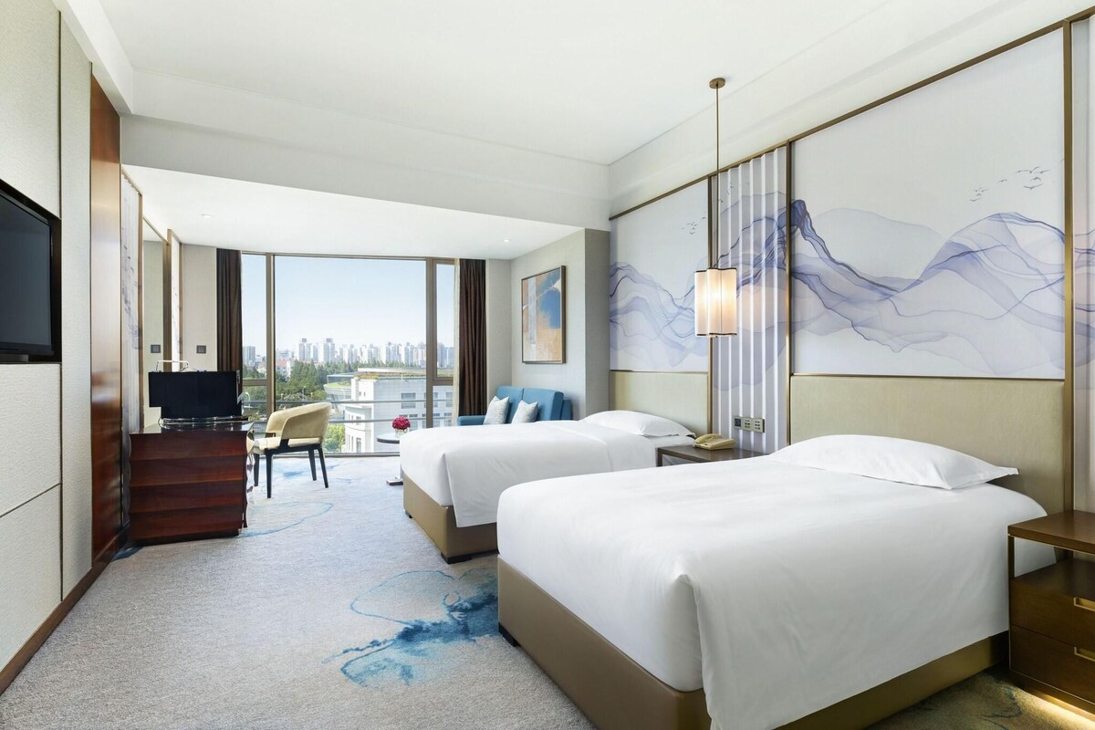A hotel room with two beds, a desk, a TV, and a large window offering a city view.