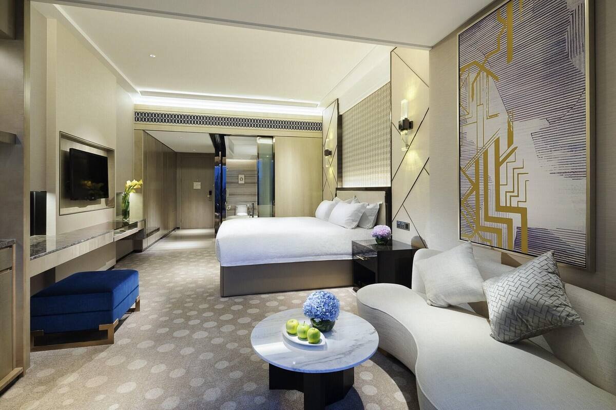 Une chambre d’hôtel moderne avec un lit, un canapé, une table basse avec des pommes et un vase, une télévision et un grand tableau abstrait.