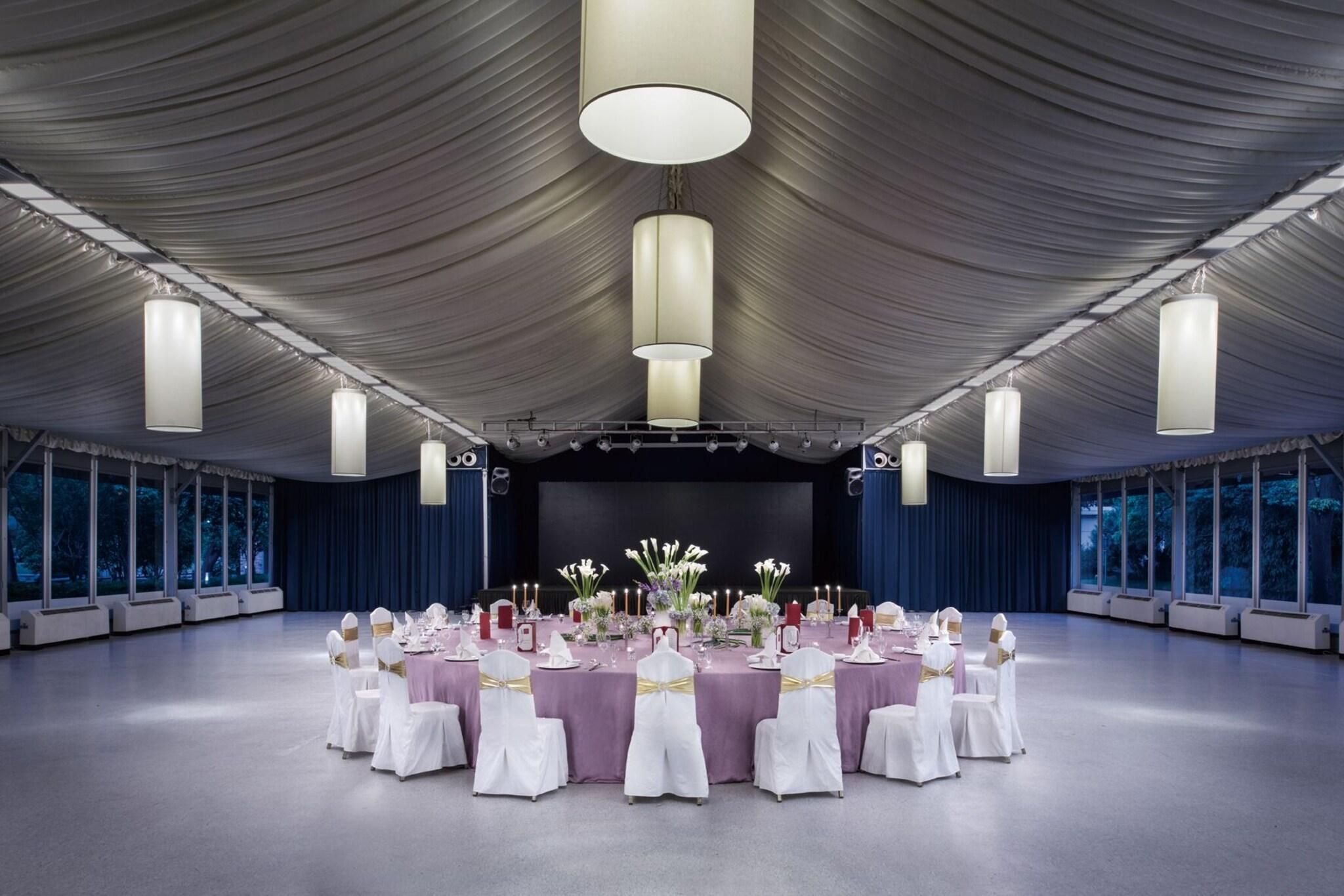 A large banquet hall with round tables, white chairs, and a stage with a backdrop.