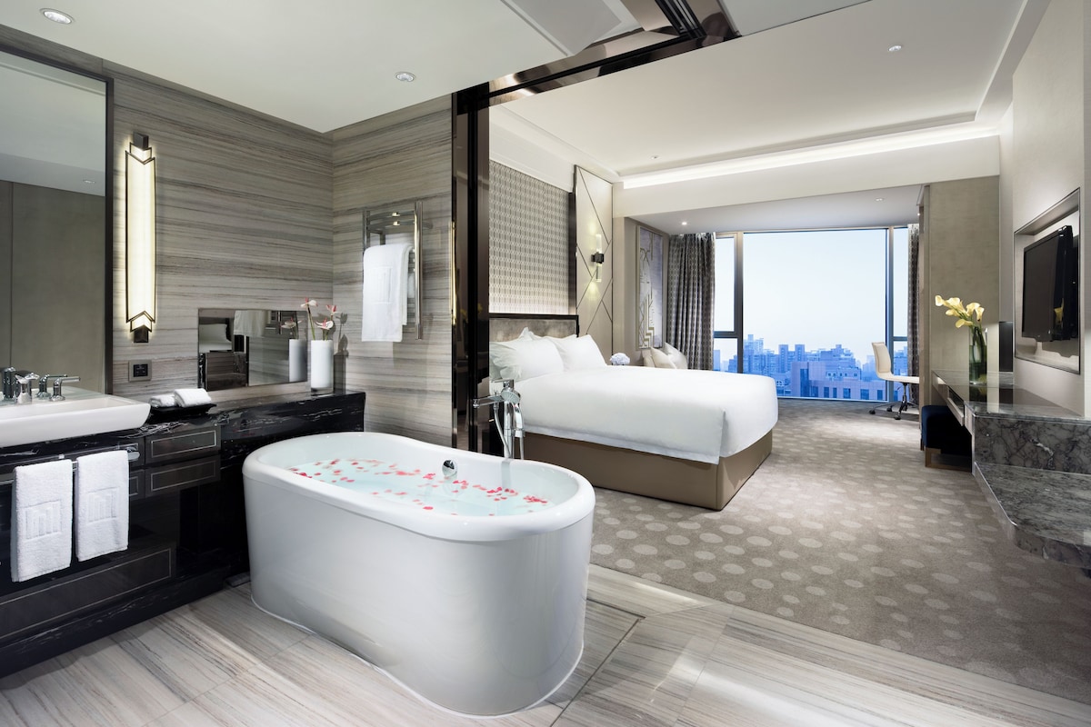 A modern hotel room with a large bathtub, a bed, a bathroom with a sink and mirror, and a city view.