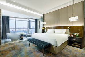 A modern hotel room with a large bed, a bathtub, a bench, a desk, and a view of the city.