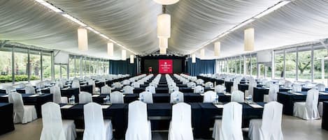 A large conference room with rows of tables and chairs, a stage with a red backdrop, and large windows.