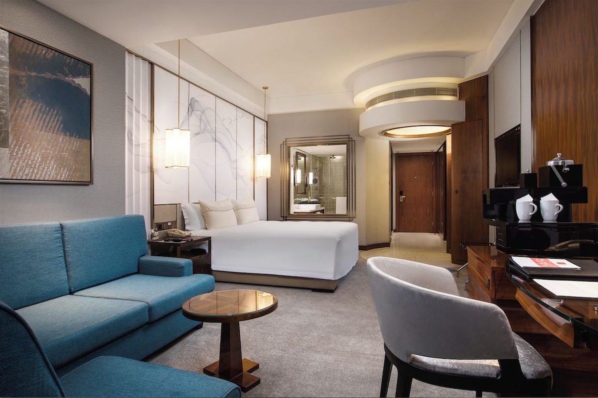 A hotel room with a bed, a blue sofa, a round table, a chair, a desk with a coffee machine, and a TV.