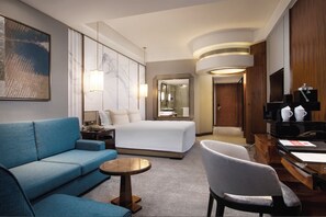 A hotel room with a bed, a blue sofa, a round table, a chair, a desk with a coffee machine, and a TV. - Grand Millennium Shanghai Hongqiao (Shanghai)
