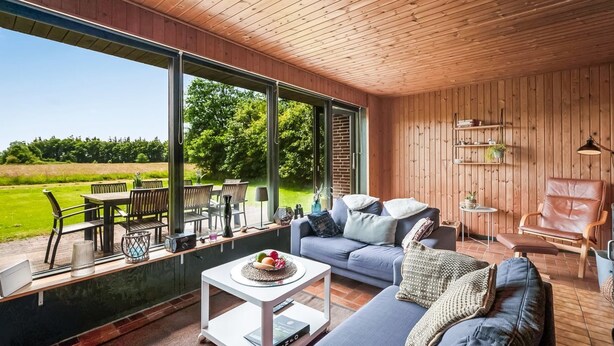 Un intérieur moderne avec un plafond en bois, de grandes fenêtres, un canapé bleu, un coin repas et une vue sur un champ verdoyant.